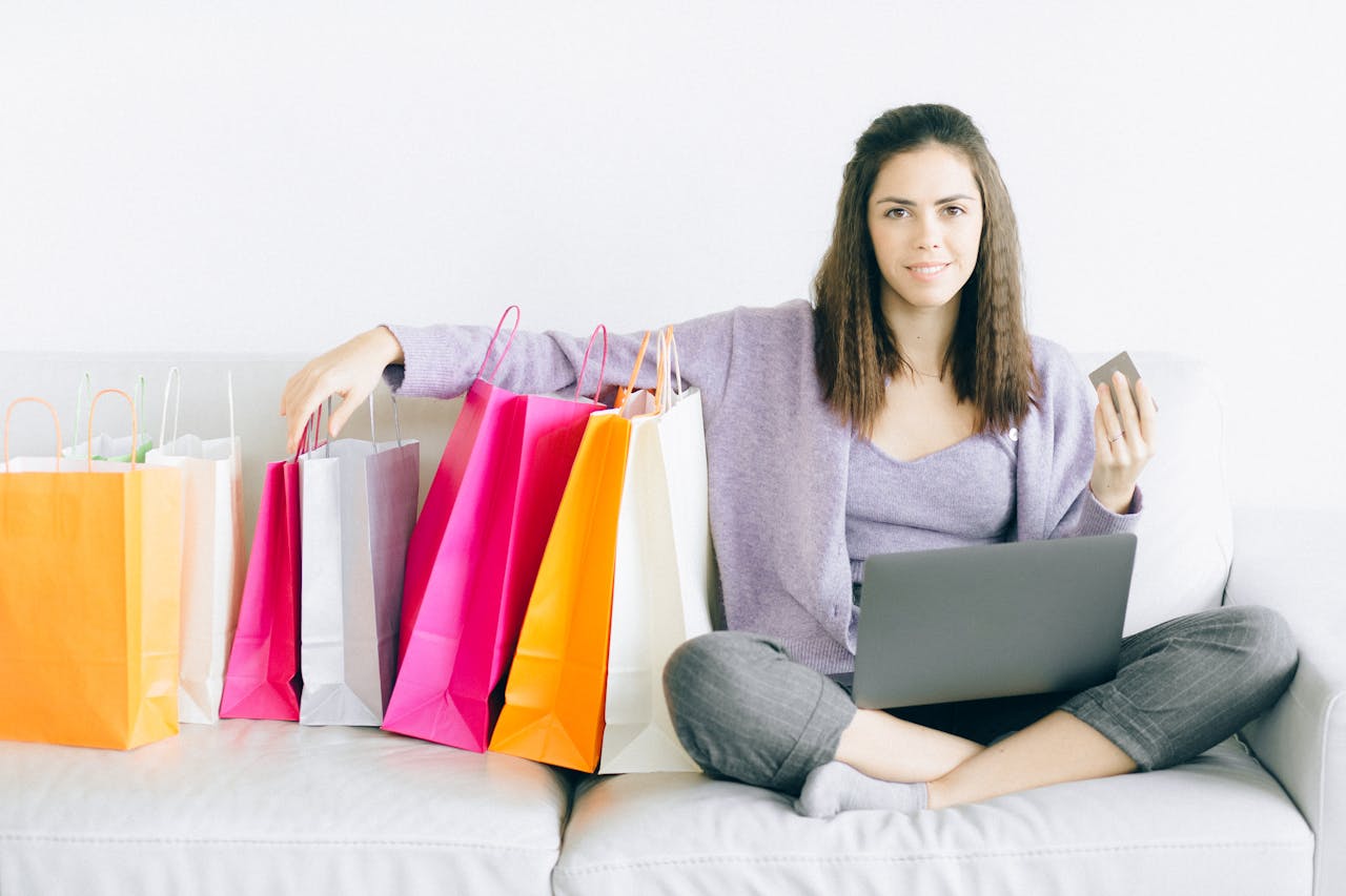 about-img Young woman enjoying online shopping with a laptop and colorful bags on a sofa.